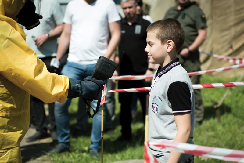 A man in a yellow hazmat suit simulates checking radiation levels on a Ukrainian boy by holding a black device out in front of him. Red-and-white striped tape keeps them separated from an onlooking crowd in the background. 