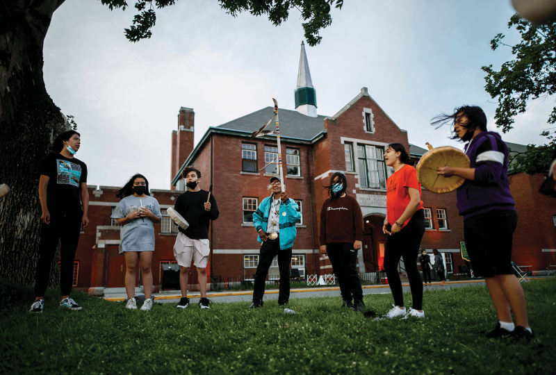A group of Indigenous elders and students stand in a circle praying in front of a red-brick residential school.