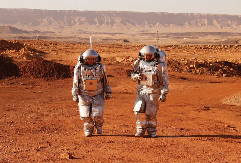 Two people in astronaut suits walk in an orange-brown desert landscape
