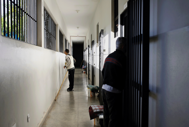 Pictured is a hallway with white walls and barred windows. There is a man in a black pants and a white sweatshirt looking out of the window, with another man standing down the hall looking at him. 