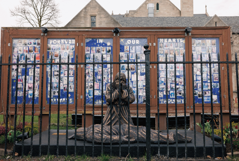 The image shows a sculpture of a weeping person kneeling over a dead body with a display in the back showing faces of people killed by gun violence. 