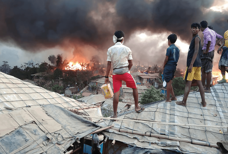 Rohingya youth stand atop tarp roofs as they watch fire spread in the treeline