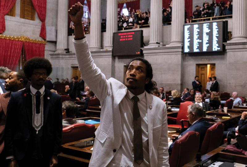 Representative Justin Jones, a Black-Filipino man in a white suit and brown tie, stands amid the aisles of Tennessee's House of Representatives, raising his fist to the ceiling as his colleague Justin Pearson stands behind him to the left.