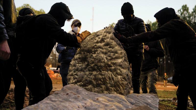 A group of protestors wearing masks, hats, and sweatshirts touch and gather around a memorial stone dedicated to Manuel Esteban “Tortuguita” Paez Terán, a 26-year-old demonstrator killed by law enforcement.