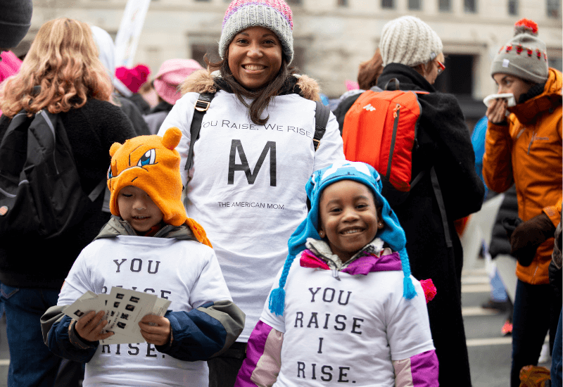 A woman and two children stand in a crowd, wearing white t-shirts that say "You Raise I Rise"