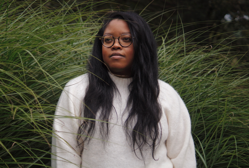 Cole Arthur Riley, a Black woman wearing glasses and a white sweater, stands before tall grasses