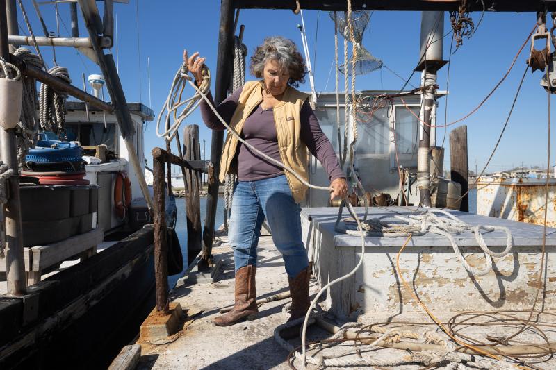 The author of this piece, Diane Wilson, is wearing a tan vest with a purple long-sleeve shirt, jeans, and brown boots. She's rolling up rope on a boat, which is docked next to another one to the left.