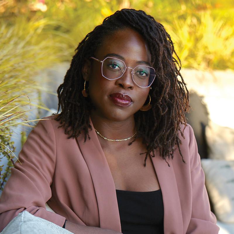 A photo of Chichi Agorom: author of 'The Enneagram for Black Liberation.' She is a black woman with long hair and glasses. She's wearing a light red jacket and black blouse as she sits outside next to a concrete wall.