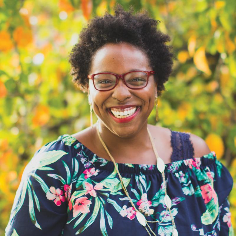 A photo of Jessica Denise Dickson: a life empowerment coach and Enneagram guide. She is a black woman with afro styled hair. She's wearing a dark blue dress with floral patterns, a long gold necklace, and red glasses. She's outside with trees behind her.