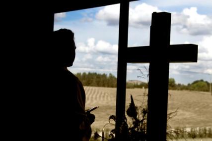 A clergy member next to a cross looking out a window. Photo courtesy Berents via Shutterstock. Via RNS.