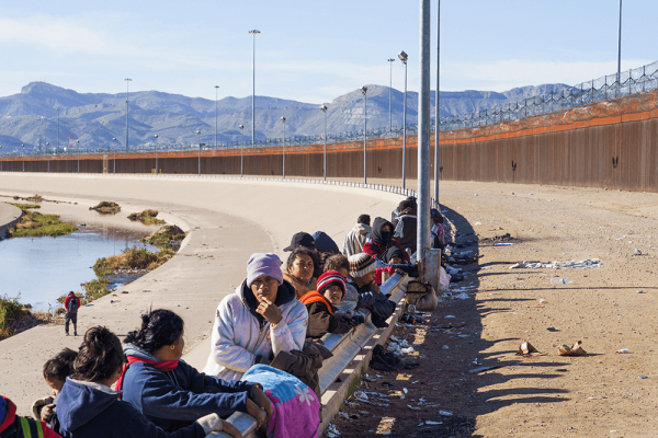 A line of people wearing hats and sweatshirts lean against a low fence next to the U.S.-Mexico border wall. Mountains are in the distance.
