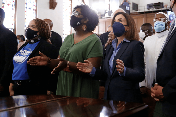 A diverse crowd of people stand in a church wearing masks during a vigil.