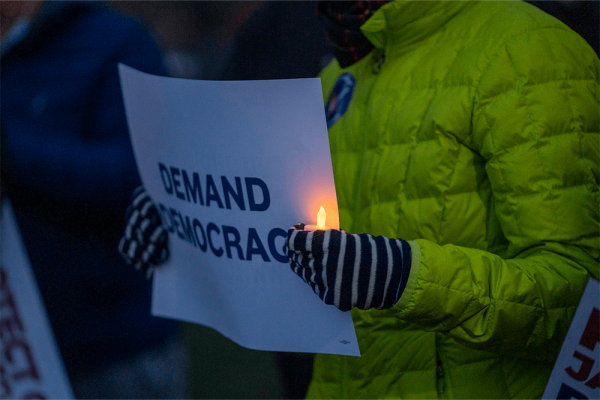 A person wearing a green puffy coat holds a lit candle and sign that says "demand democracy"