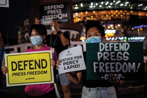 A group of Filipinos stand together, wearing masks and carrying signs that hold variations on the phrase "Defend Press Freedom"