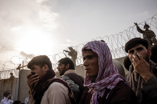 Afghan men stand before a concrete wall topped with barbed wire and a U.S. soldier