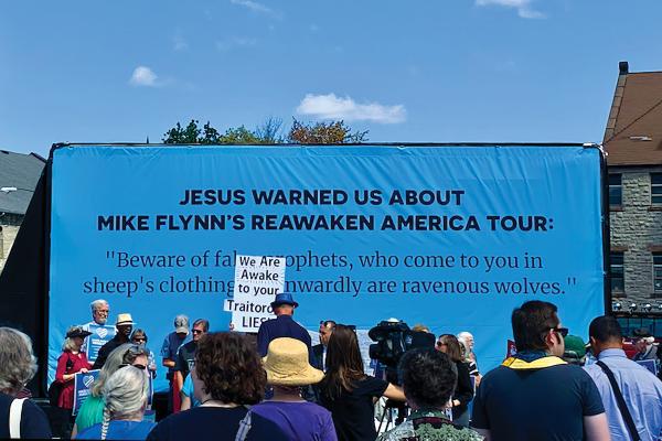 Photo of a cerulean blue billboard with black text that reads "Jesus Warned Us About Mike Flynn's ReAwaken America Tour." Foreground features a group of people facing the billboard; one protestor's sign reads "We Are Awake to Your Traitorous Lies"