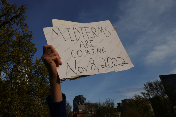 A demonstrator holds a sign reading "Midterms Are Coming Nov 8, 2022" against a blue sky.