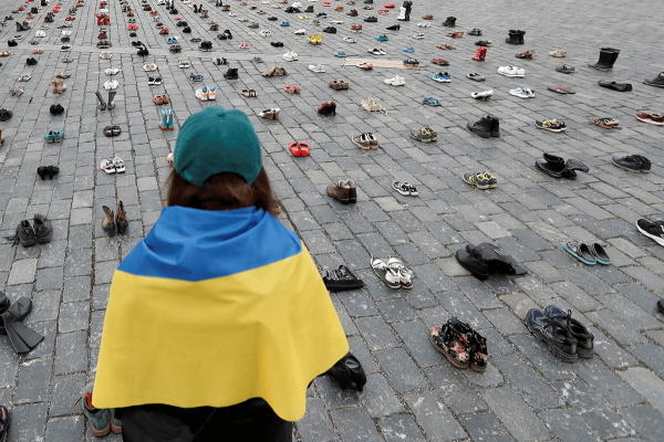 A woman wrapped in a Ukrainian flag looks at pairs of shoes arranged in rows.