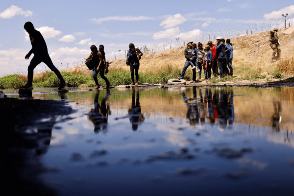 Shadows fall over migrants who are crossing a river. Their reflections appear in the surface.