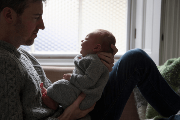 A man holds a crying baby in front of a window.