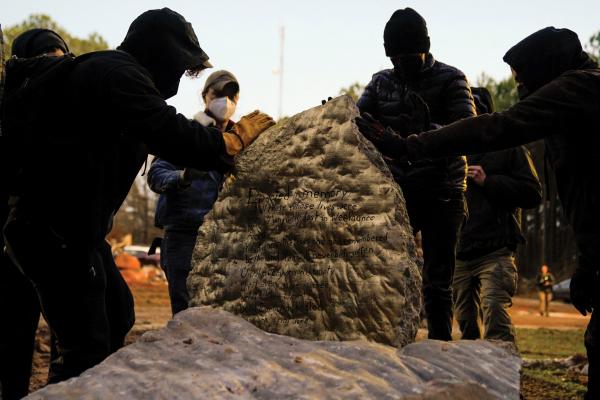 A group of protestors wearing masks, hats, and sweatshirts touch and gather around a memorial stone dedicated to Manuel Esteban “Tortuguita” Paez Terán, a 26-year-old demonstrator killed by law enforcement.