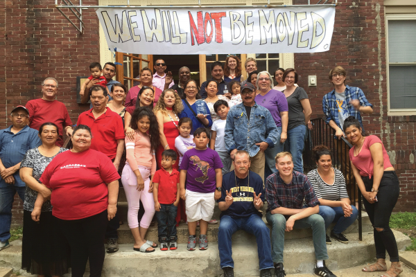 The photo shows a large group of people smiling for a photo underneath a banner that reads "We Will Not Be Moved." The people are standing in front of the door to their apartment complex. 