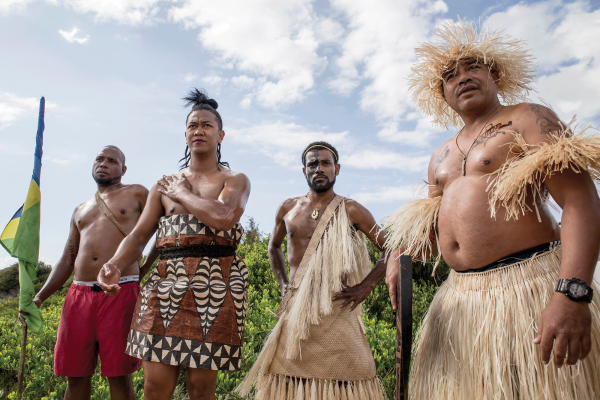 The photo shows four men from the Pacific Islands, some are in traditional dress made of straw and other natural fibers, and one of the men is holding a flag that is wrapped around the flag pole