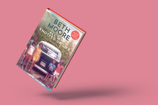 An image of Beth Moore's memoir 'All My Knotted-Up Life' against a pink backdrop. The cover shows her as a child posing with her family as they stand behind and to both sides of an old navy and white van outside near a forest.