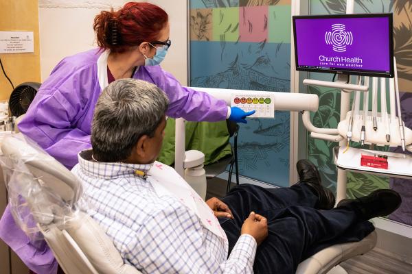 A side-rear view woman doctor with red hair points to a screen with a spectrum of faces from sad to happy, asking her patient in the chair (a man with gray hair) which is most accurate for him. A purple screen with "Church Health" is shown nearby.