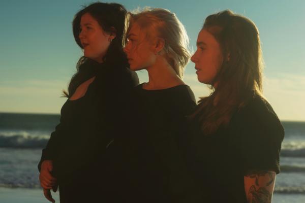 From left to right, musicians Lucy Dacus, Phoebe Bridgers, and Julien Baker are dressed in black and cast in the warm glow. They stand in a cascading line next to one another, staring off beyond the left side of the photo with waves in the background.