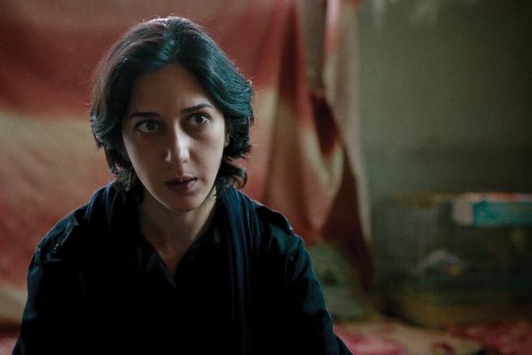 A photo of actress Zar Amir Ebrahimi as fictional journalist Arezoo Ramimi in the film 'Holy Spider.' She is cast against a red flag in the background and staring just off camera at something.