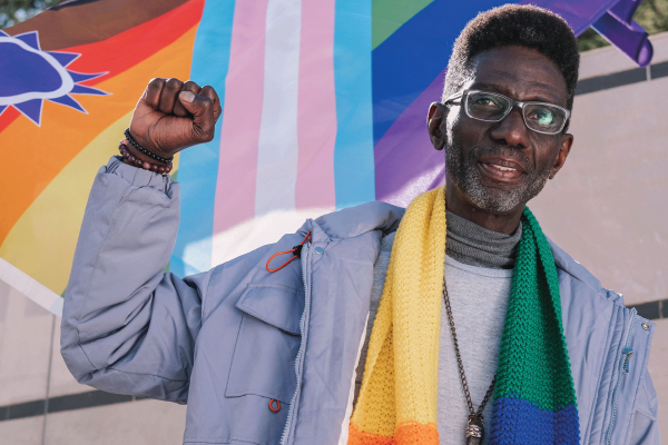 The image shows an older Black man wearing a knitted rainbow scarf and holding up a fist in front of a colorful pride flag. 