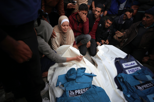 The photo shows mourning Gazans as they stand over the covered bodies of two slain journalists, their blue press vests resting on their bodies. 