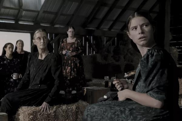 A group of Mennonite women are standing and sitting in a barn filled with crates and hay bales in the film 'Women Talking.'