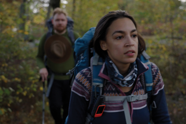 Foreground of image shows Rep. Alexandria Ocasio-Cortez hiking in a green forest. She wears a geometric sweater and a blue backpack; her boyfriend is pictured hiking behind her, slightly out of focus