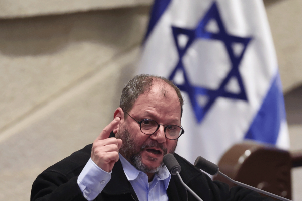 The image shows an older man in a suit and glasses holding up a finger emphatically while speaking in to a microphone. There is an Israeli flag behind him. 
