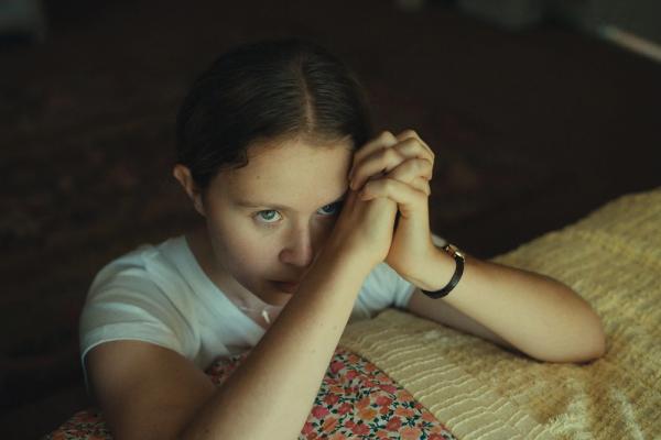 A young white teenage girl named Jem Starling (played by actress Eliza Scanlen) is sitting on the edge of a bed. Here elbows rest on the quilt blanket with her hands folded in prayer as she looks beyond the frame toward an unseen ceiling.