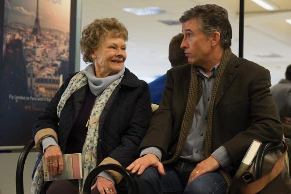 Philomena (played by Dame Judi Dench) and Sixsmith (played by Steve Coogan) sit next to each other in a waiting room. Philomena is wearing a black jacket with a flower-patterned scarf. Sixsmith is wearing a dark brown jacket and blue jeans.