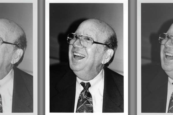 The image shows a black and white photo of a older white man laughing. He is bald and wearing glasses and a suit and tie.