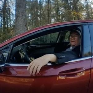 A man wearing a clergy collar sits in a red car, with his left hand out the rolled down car window.