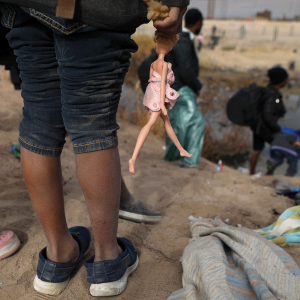 The picture shows the legs of a girl standing on the bank of the Rio Grande, holding a Barbie doll by its hair. 