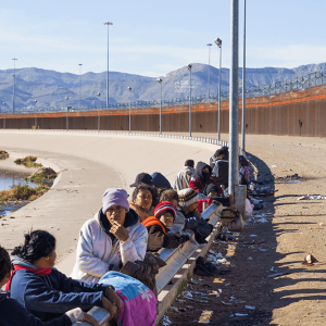 A line of people wearing hats and sweatshirts lean against a low fence next to the U.S.-Mexico border wall. Mountains are in the distance.