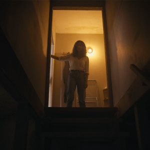 A back-lit woman stands at the top of basement stairs, looking down.