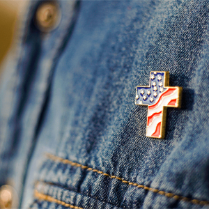 A close up of Christian cross pin with American flag colors is pinned on blue jeans jacket. 