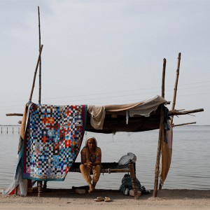 A woman sits on a bench under a makeshift shelter made of sticks and a quilt. Behind the shelter, floodwaters stretch to the horizon.