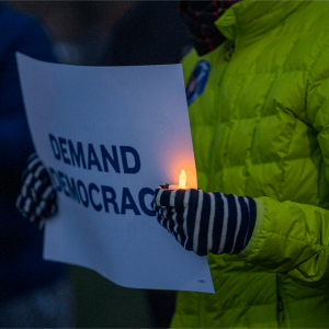 A person wearing a green puffy coat holds a lit candle and sign that says "demand democracy"