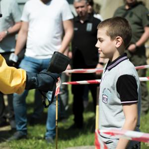 A man in a yellow hazmat suit simulates checking radiation levels on a Ukrainian boy by holding a black device out in front of him. Red-and-white striped tape keeps them separated from an onlooking crowd in the background. 