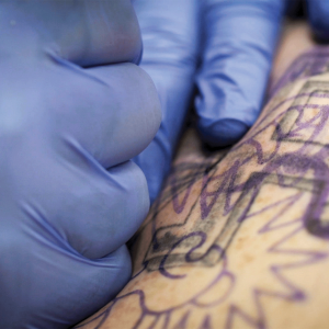 The photo shows two hands wearing blue latex gloves tattooing over a tattoo of a swastika 