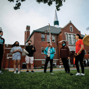 A group of Indigenous elders and students stand in a circle praying in front of a red-brick residential school.