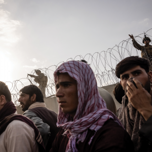 Afghan men stand before a concrete wall topped with barbed wire and a U.S. soldier